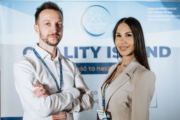 Two professionals posing at a conference booth with blue banners, wearing lanyards and name badges.