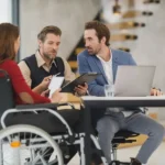 Zespół w biurze omawia dokumenty przy laptopie, jedna z osób na wózku, dostępność cyfrowa w zamówieniach publicznych